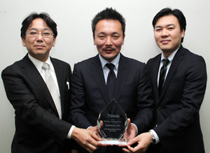 ヘルスケアIT事業部 部長　田中　英敏 氏（中） 開発2課リーダー　宮崎　秀隆 氏（右） マーケティングセールス課　長谷川　篤 氏（左） FileMaker Excellence Awardのトロフィーとともに。