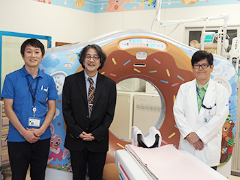 子ども向けにペインティングされた64列CT「Discovery CT 750 HD」（GEヘルスケア社製）と，左から放射線診療部の永松洋志特殊撮影主任，宮嵜診療部長，鈴木成人技師長