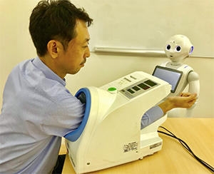 Pepperによる血圧測定イメージ （顔認識でリラックス度を判定。測定器はPepperと自動連動）